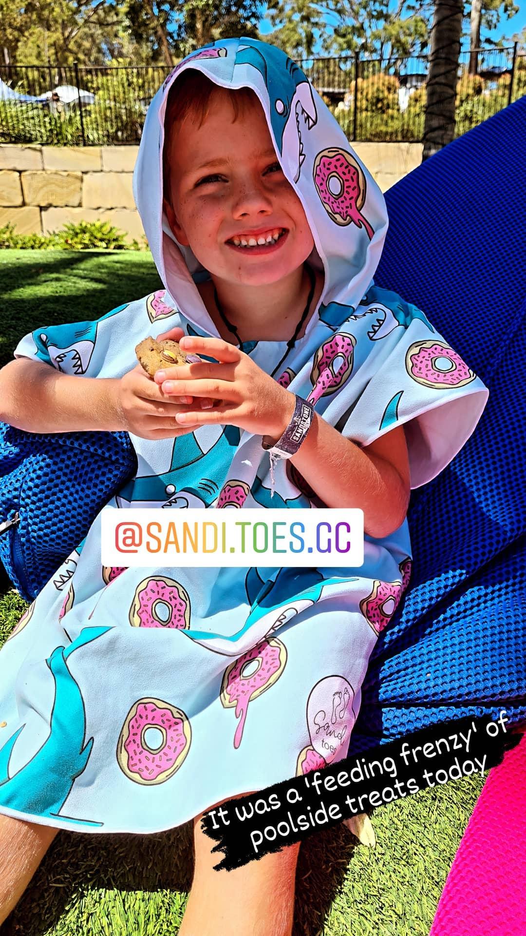 Here we have a boy sitting eating his lunch wearing his Feeding Frenzy sand less hooded towels, the designs are of sharks and donuts on a blue back ground