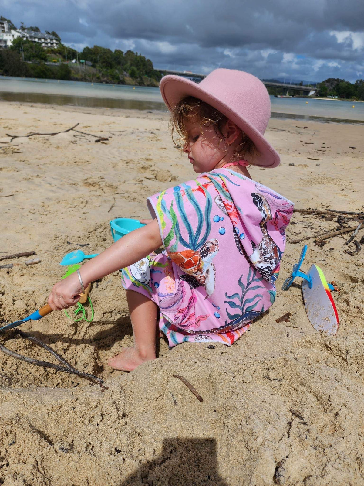 Young Girl sitting on a beach in Queensland with her Turtle Cove sand less hooded beach towel, here she is wearing a wide brim pink hat which compliments the pink turtle cove print 