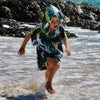 Boy (ages 6-12) wearing a blue hooded beach hoodie with a Bird of Paradise design featuring a yellow crested white cockatoo and palm leaves. He is running happily through the ocean at Currumbin Beach, Queensland, with rocks visible in the background.