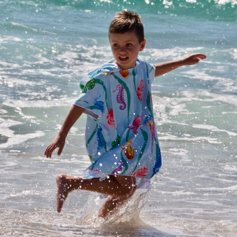 Boy (ages 6-12) wearing a [hoodie color] Earth & Sea hooded beach hoodie. The design features a vibrant underwater scene teeming with colorful fish, a curious chameleon, and a friendly sea horse.